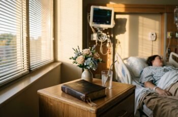 Exemplar da Bíblia Sagrada sobre a mesa de cabeceira de um quarto hospitalar ao lado de flores sob luz suave matinal